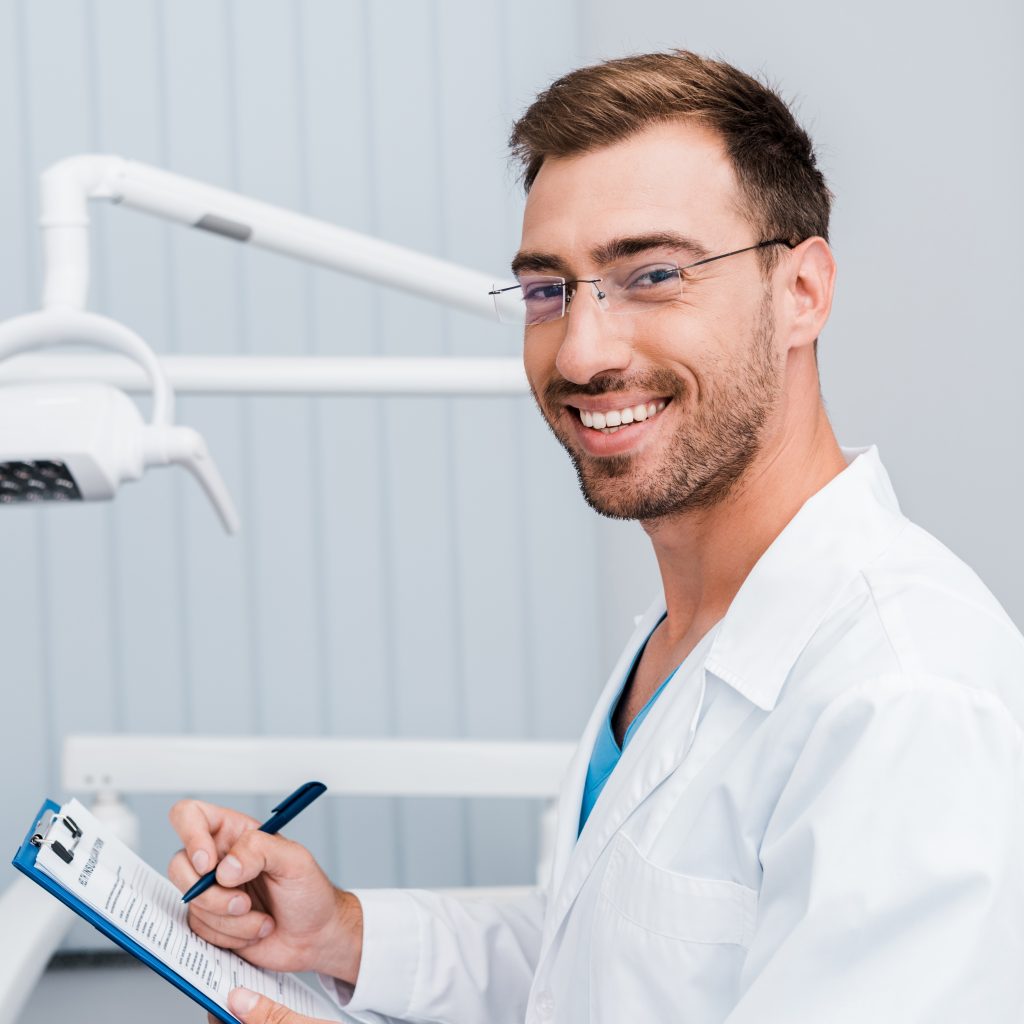 happy bearded man in white coat and glasses holding pen and clipboard in dental clinic